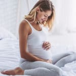 Eine schwangere Frau sitzt auf dem Bett und legt die Hände auf den Bauch