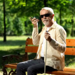 Ein Sehbehinderter Mann sitzt auf einer Parkbank und nutzt sein Smartphone